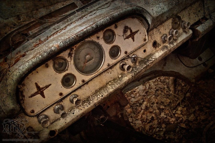 A close-up, diagonal view of a rusted and weathered dashboard from an abandoned 1957 Ford. The off-white metal instrument panel features circular gauges and two rusted four-pointed star accents, surrounded by peeling paint, rust, and dirt, with dry forest leaves visible underneath.